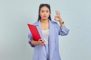 retrato de uma jovem empresária asiática positiva de terno segurando a pasta de documentos e faz um gesto bem, concorda com a boa ideia isolada no fundo roxo foto