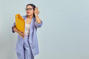 retrato de uma jovem empresária asiática séria mantém a mão perto da orelha tenta ouvir conversa privada ouve fofocas enquanto segura pasta amarela isolada no fundo roxo