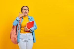 estudante asiática jovem chata cansada em roupas jeans com mochila segurando o notebook isolado em fundo amarelo. conceito de estilo de vida islâmico religioso de pessoas foto