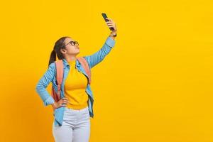 retrato de estudante asiática jovem confusa em roupas casuais com mochila usando smartphone e procurando rede de internet isolada em fundo amarelo. educação no conceito de universidade universitária foto