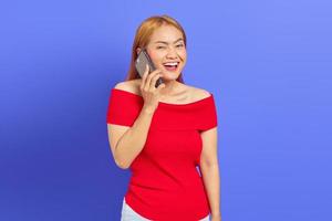 retrato de uma jovem asiática feliz em vestido vermelho, regozijando-se enquanto fala no celular isolado no fundo roxo foto