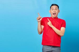 retrato de jovem asiático surpreso na camisa vermelha em pé apontando o dedo no espaço de cópia isolado no fundo azul