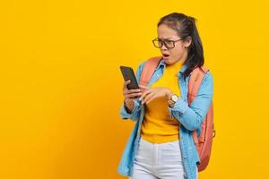 retrato de estudante asiática jovem surpresa em roupas casuais com mochila digitando mensagem de texto ou rolando feed na rede social usando smartphone isolado em fundo amarelo foto