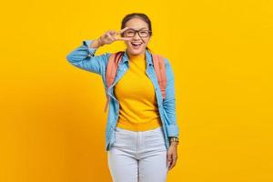 alegre jovem estudante asiática em roupa jeans com mochila cobrindo o olho com gesto de vitória v-sign isolado em fundo amarelo. educação no conceito de universidade foto