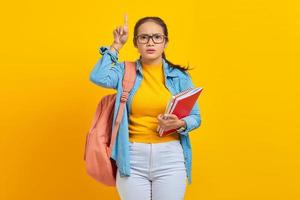 retrato de estudante asiática jovem pensativa em roupas jeans com mochila segurando o caderno e apontando para cima com o dedo sobre fundo amarelo. educação no conceito de faculdade universitária do ensino médio foto