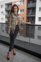 linda mulher morena glamour com jaqueta xadrez posando na varanda moderna com vista incrível sobre a cidade retrato de uma mulher elegante elegante com pernas longas, calça jeans preta vestindo na varanda foto