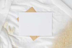 maquete de convite de cartão em branco no envelope marrom com grama seca de rabos de coelho no fundo branco, composição mínima do local de trabalho da mesa, postura plana, maquete foto