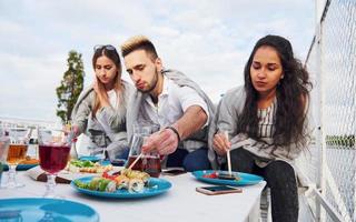 felizes jovens amigos estavam sentados em uma mesa e fazendo um piquenique ao ar livre foto