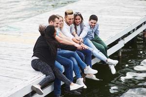 retrato de alegre grupo de jovens sentados à beira do cais, ao ar livre na natureza. amigos curtindo um dia no lago. foto