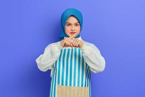 retrato de mulher jovem dona de casa séria em hijab e avental listrado, dedo cruzado e mostrando o gesto de recusa isolado no fundo roxo. conceito de estilo de vida muçulmano dona de casa de pessoas foto