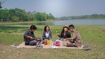 grupo de jovens mulheres e homens sentados, comendo e tocando violão no parque na primavera verão foto