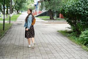 uma garota de vestido e uma jaqueta jeans com uma mochila laranja nos ombros vai para a escola e olha para trás. conceito de volta às aulas. fotografia de rua. foto