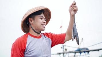 feliz jovem pescador na praia segurando seu peixe e mostra na frente de seu barco foto