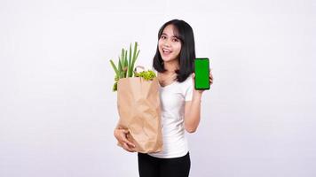 mulher asiática com saco de papel de legumes frescos e segurando um telefone de tela verde com fundo branco isolado foto