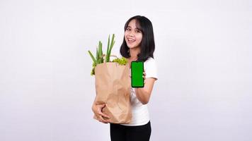 mulher asiática com saco de papel de legumes frescos e segurando um telefone de tela verde com fundo branco isolado foto