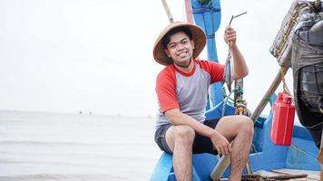 feliz jovem pescador na praia segurando seu peixe e mostra na frente de seu barco foto