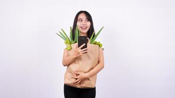 mulher asiática com saco de papel de legumes frescos e segurando um telefone com fundo branco isolado foto