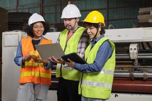 trabalhadores de uniformes de segurança e engenheiros industriais com capacete de segurança usam computador portátil para verificar e controlar máquinas. três profissionais trabalham na fábrica de fabricação de papel, mantêm equipamentos de produção. foto