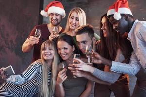 grupo de jovens lindos fazendo selfie na festa de ano novo, melhores amigos, meninas e meninos juntos, se divertindo, posando de pessoas de estilo de vida emocional. chapéus de Papai Noel e taças de champanhe nas mãos foto