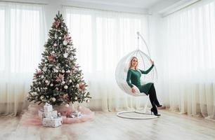feliz Natal e Feliz Ano Novo. linda mulher loira em um vestido verde sentado na cadeira suspensa na árvore foto