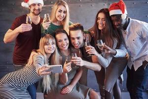 grupo de jovens lindos fazendo selfie na festa de ano novo, melhores amigos, meninas e meninos juntos, se divertindo, posando de pessoas de estilo de vida emocional. chapéus de Papai Noel e taças de champanhe nas mãos foto