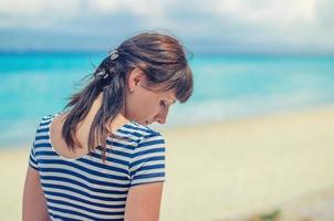 menina bonita com camisa listrada e cabelo escuro, posando e olhando para baixo na praia foto