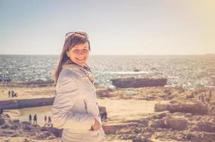 viajante jovem com jaqueta branca e óculos de sol, olhando para a câmera e sorrir na costa da baía de dwejra foto