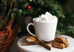 café em uma xícara branca com marshmallows. café festivo da manhã com biscoitos de amêndoa cantuccini italianos tradicionais. uma xícara de café em um fundo de ramos de abeto verde em um carrinho branco. foto