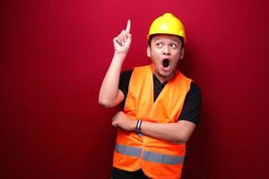 choque jovem trabalhador asiático está surpreso e gritando uau com apontando acima com a mão isolada no fundo vermelho. foto