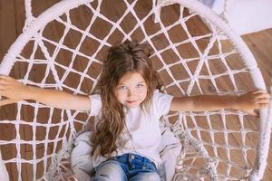 adorável menina alegre em t-shirt branca e jeans em elegante cadeira de balanço interior. moda, estilo, infância, emoções, conceito de crescimento foto