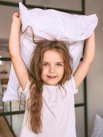 adorável menina alegre no travesseiro de luta de t-shirt branca em casa interior. moda, diversão, estilo, infância, emoções, conceito de crescimento foto
