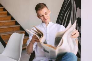 homem adolescente moderno lendo jornal de papel durante o café da manhã em casa. leitura, moda antiga, moderna, conceito de notícias foto