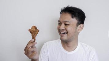 jovem asiático está comendo frango frito, vestindo camisa branca, sentindo-se feliz e sorrindo foto