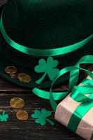 chapéu de duende, moedas de ouro, trevo de trevo e presente de fita verde sobre fundo escuro de madeira. símbolos de boa sorte para st. dia de patrício. foto