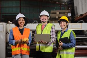 trabalhadores de uniformes de segurança e engenheiros industriais com capacete de segurança usam computador portátil para verificar e controlar máquinas. três profissionais trabalham na fábrica de fabricação de papel, mantêm equipamentos de produção. foto