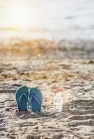 chinelos na areia da praia com água em garrafa plástica e raio de sol foto