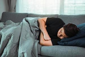 homem asiático sente a doença enquanto estava com frio deitado no sofá e vestindo um cobertor em sua casa. foto