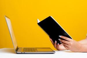 mão e livro vista lateral lendo em uma mesa branca há um laptop na frente. conceito de leitura de livro relaxante lendo o pano de fundo amarelo foto