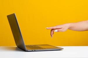 mãos e laptop de uma jovem que está determinada a trabalhar nos recados on-line mão apontando para o computador na frente wan no pano de fundo amarelo da mesa branca foto