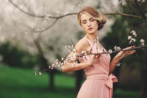 jovem elegante perto de árvore florescendo no parque. fundo de primavera. loira com penteado de vestido rosa. copie o espaço. foto