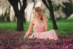 bela jovem loira em flores coloridas. garota com maquiagem e penteado de vestido rosa no parque primavera florescendo. obra de arte da mulher romântica. modelo de ternura bonita olhando para a câmera foto