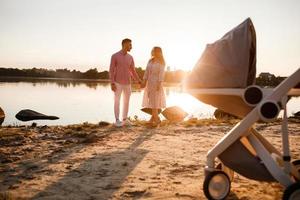 lindo jovem casal apaixonado com bebê andando no lago ao pôr do sol. carrinho de bebê no fundo frontal. família tem um bom tempo juntos. espaço de cópia foto