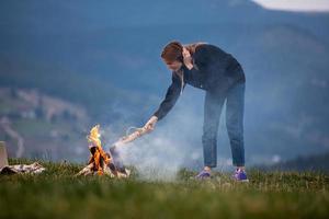 jovem freelancer joga lenha em um incêndio nas montanhas à noite. garota turista descansando e trabalhando ao ar livre foto