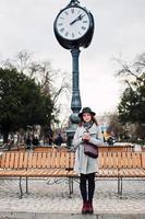 menina modelo jovem em um casaco cinza e chapéu preto com bolsa de couro nos ombros ficar com copo de plástico de fundo de café velho relógio vintage na rua da cidade. foto