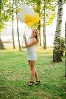 retrato de menina loira vestindo vestido branco com balões nas mãos contra o parque na festa de despedida.