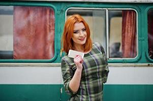 senhora jovem ruiva com telefone celular móvel e fones de ouvido, vestindo no vestido xadrez fundo velho ônibus de minivan turquesa vintage. foto