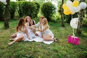 cinco garotas com champanhe na mão vestidas de vestido branco na despedida de solteira sentado ao ar livre no parque e se divertindo. foto