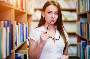 menina morena na biblioteca com óculos na mão, use blusa branca e mini saia preta. mulher de negócios sexy ou conceito de professor. foto