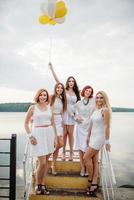 cinco meninas com balões na mão usavam vestidos brancos na festa de despedida contra o cais no lago. foto
