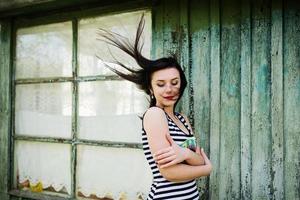 menina modelo morena com cabelo no ar usando vestido com listras fundo ciana casa de madeira com janelas antigas. foto